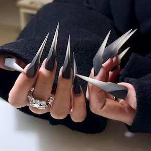 May include: Black, long, pointed acrylic nails with a gradient design, transitioning from black to gray. The nails are adorned with silver-colored, geometric nail art. A silver ring with clear stones is also visible.