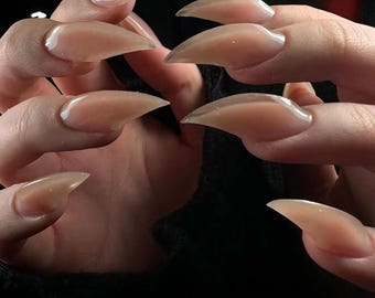 May include: Close-up of hands with long, almond-shaped, nude-colored acrylic nails. The nails are glossy and have a sharp, pointed tip. The background is dark, emphasizing the nails' shape and color.