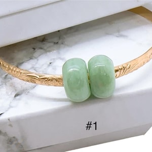 May include: A gold-colored bangle bracelet with an engraved floral pattern. Two light green, oval-shaped beads are centered on the bracelet. The number "#1" is printed in black on the white surface below the bracelet.