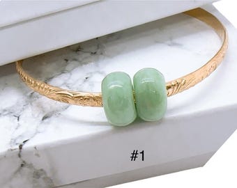 May include: A gold-colored bangle bracelet with an engraved floral pattern. Two light green, oval-shaped beads are centered on the bracelet. The number "#1" is printed in black on the white surface below the bracelet.