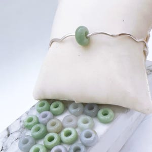 May include: A silver bracelet with a wavy design, featuring a single, round, light green jade bead. Several loose jade beads in varying shades of green and lavender are scattered below. The beads have a donut shape.