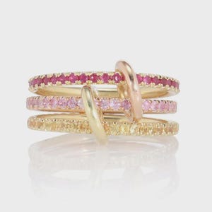 Könnte beinhalten: Vier goldene Ringe, die übereinander gestapelt sind, jeder mit einer Reihe kleiner Edelsteine in Rot-, Rosa- und Gelbtönen. Zwei gebogene Goldbänder umschließen die Ringe und schaffen ein einzigartiges Design. Die Ringe werden auf einer reflektierenden Oberfläche präsentiert.