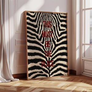 May include: A framed art print with a zebra pattern and the red text "THIS MUST BE THE PLACE". The artwork is in a gold frame, leaning against a white wall. The room has wooden flooring.