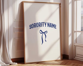 May include: A framed print featuring the text "SORORITY NAME" in navy blue, curved above a navy blue bow illustration. The print has a cream background and a gold frame, leaning against a wall with a wooden floor.