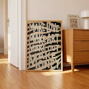 May include: A rectangular framed wall art piece featuring a black and white zebra stripe pattern background. The text "MAKE LOVE MAKE ART MAKE OUT MAKE MONEY" is printed in large, bold, off-white capital letters across the centre of the print.