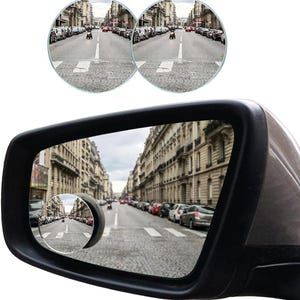 May include: A black car side mirror with two small, round blind spot mirrors attached. The mirrors reflect a city street with parked cars and buildings. The main mirror also reflects the street scene, providing a wider view.