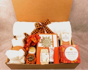 May include: A gift box filled with fall-themed items. Includes a white towel, orange and black plaid bow, decorative pumpkins, a candle, a box of chocolates, a box of tea, and a greeting card with a gold snowflake.
