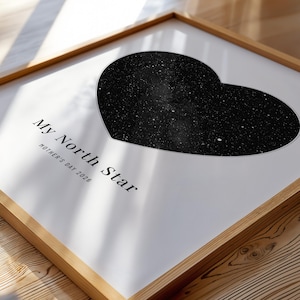 May include: Framed print featuring a black heart-shaped constellation against a white background. The text "My North Star" and "Mother's Day 2026" are printed below the heart. The frame is made of light-colored wood, resting on a wooden surface.