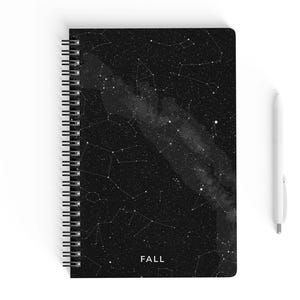 May include: A spiral-bound notebook with a black cover featuring a white constellation design. The word "FALL" is printed in white at the bottom. A white pen rests beside the notebook, creating a contrast against the dark background.