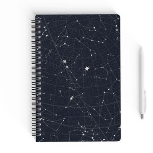May include: Spiral-bound notebook with a dark blue cover featuring a white constellation design. A white pen rests beside the notebook. The notebook's pages are blank, ready for notes or sketches.
