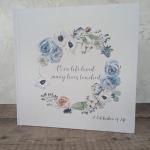 May include: A white book featuring a floral wreath in blue, white, and peach hues. The text reads "One life lived, many lives touched" with "A Celebration of Life" beneath. The book is displayed on a wooden surface.