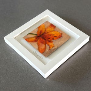 May include: A framed painting of an orange flower with dark accents. The artwork is set within a white, square frame. The artist's signature and the year 2026 are visible in the lower left corner of the frame.