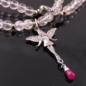 May include: A necklace with clear, round beads and a silver fairy pendant. The fairy pendant has detailed wings and a dangling teardrop-shaped, pink gemstone. The necklace is a delicate piece of jewellery.