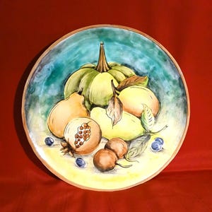 May include: Hand-painted ceramic plate featuring a still life of fruits. The plate has a light brown rim and a turquoise and yellow background. The fruits include a green pumpkin, pears, and a pomegranate. The plate is a decorative piece.