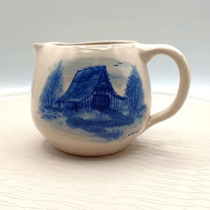 May include: Cream-colored ceramic creamer with a blue barn scene. The creamer has a rounded body, a handle, and a spout. The barn is painted in blue with trees and a small bird in the sky. The creamer is a vintage piece.