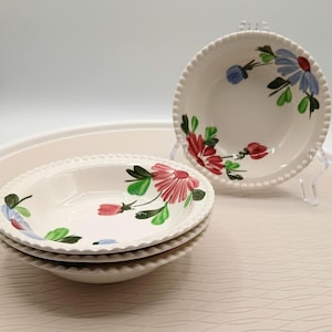 May include: A stack of three white ceramic bowls with a floral design. The bowls have a beaded edge and are decorated with red, pink, blue, and green flowers. One bowl is displayed on a clear stand.