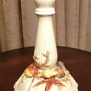 May include: White ceramic candle holder with a seashell design. The holder features a starfish, seashells, and coral.
