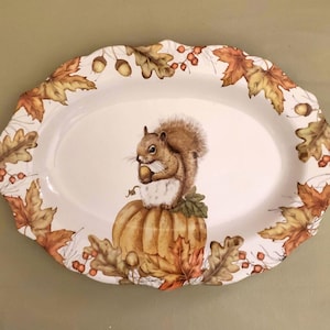 May include: White oval serving platter with a brown squirrel holding an acorn sitting on top of a brown pumpkin. The platter is decorated with fall leaves in shades of orange, red, and yellow.
