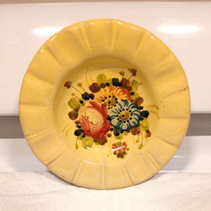 May include: A yellow ceramic ashtray with a floral design. The flowers are painted in shades of red, blue, yellow, and green.