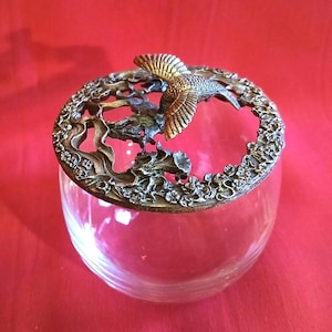 May include: A clear glass jar with a metal lid featuring a detailed hummingbird design. The lid has a floral pattern and a cutout of a hummingbird in flight.