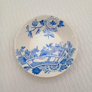 May include: A vintage, off-white ceramic plate with a blue floral and landscape design. The rim features a bird emblem. The plate's design includes flowers, leaves, and a detailed scene of a cart.