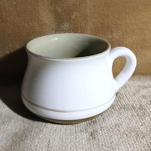 May include: A white ceramic mug with a brown rim and a wide, rounded shape. The mug has a single handle.