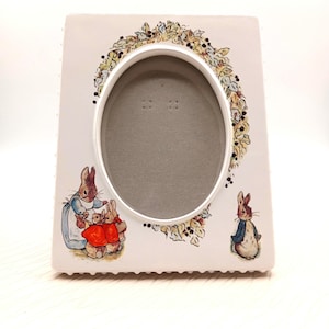May include: White ceramic picture frame with an oval opening, decorated with illustrations of rabbits and a floral wreath. The frame has a scalloped edge and is suitable for a 4x6 inch photo.
