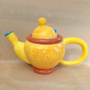 May include: A whimsical yellow ceramic teapot with a rounded body, a brown ring around the middle, and a curved yellow handle. The spout has a blue tip, and the lid is topped with a yellow knob. The teapot sits on a red base.