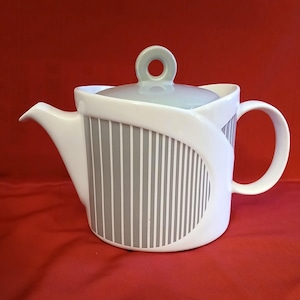 May include: White ceramic teapot with a gray and white striped design on the side. The teapot has a curved handle and a spout. The lid is light gray with a ring-shaped handle. The teapot is on a red surface.