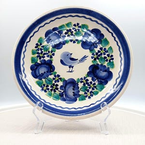 May include: A ceramic plate with a blue bird and floral design. The plate features a white base with a blue border and a central design of blue flowers and green leaves. The bird is light blue. The plate is displayed on a clear stand.