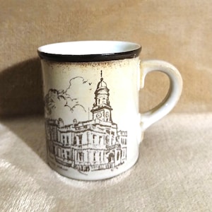May include: A white ceramic mug with a brown rim and a black and white illustration of a building with a clock tower. The building is surrounded by trees and a cloud.