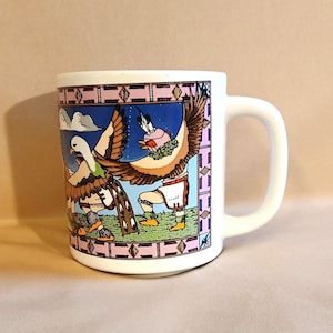 May include: White ceramic mug with a colorful design featuring a Native American figure with two birds, one white and one green, against a blue sky with clouds. The design is framed with a pink, blue, and brown geometric border.
