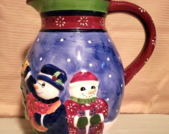 May include: A ceramic pitcher with a winter theme. The pitcher features a blue background with white snowflakes and a painted scene of two snowmen. The handle is burgundy with white accents, and the rim is green and red.