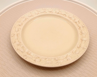 May include: A cream-colored ceramic plate with a decorative embossed border featuring floral and leaf patterns. The plate is centered on a light pink surface with a subtle geometric design, creating a classic aesthetic.
