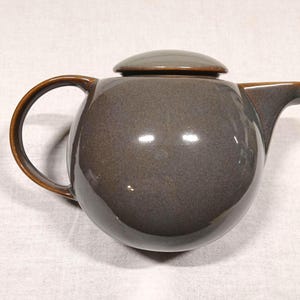 May include: A dark gray ceramic teapot with a brown handle and lid. The teapot has a rounded body and a spout on the side.