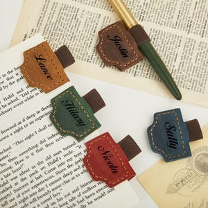 Op de afbeelding: Leren bladwijzers in verschillende kleuren: bruin, groen, rood en blauw. Elke bladwijzer heeft een naam: Lance, Hilary, Nicola en Sally. Een bruine bladwijzer met de naam Justin ligt bij een pen op een pagina.