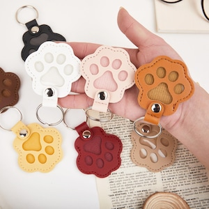 May include: A collection of paw-shaped keychains in various colors, including white, pink, orange, yellow, red, brown, and black. Each keychain features a metal ring and a leather strap for attachment.