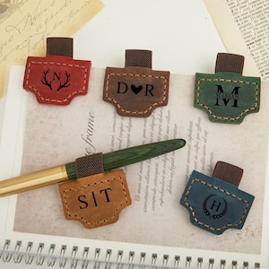 Könnte beinhalten: Eine Sammlung personalisierter Leder-Lesezeichen in Rot, Braun, Grün, Beige und Blau. Jedes Lesezeichen zeigt ein individuelles Initialen- oder Monogramm-Design mit einer braunen elastischen Schlaufe. Ein Stift liegt auf einem Notizbuch, mit dem Wort "frame".