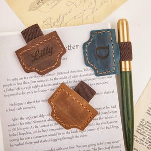 Op de afbeelding: Leren bladwijzers in bruin en blauwgroen met gestikte randen. Eén bladwijzer heeft de naam "Billy" gegraveerd, een andere de letter "D". Een groene pen met een gouden punt is ook zichtbaar.
