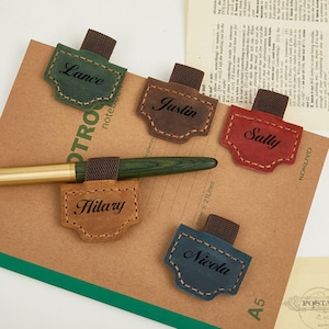 May include: A collection of personalized leather pen holders in various colors: green, brown, red, tan, and blue. Each holder is labeled with a name in black script. A green pen with a gold-colored tip rests on a brown notebook.