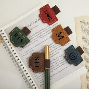 Puede incluir: Varios sujetadores de bolígrafo de cuero en diferentes colores, cada uno con una inicial o monograma único. Están unidos a un cuaderno y a un bolígrafo, presentando un tema de papelería personalizada. Los colores incluyen rojo, verde, marrón, beige y azul.