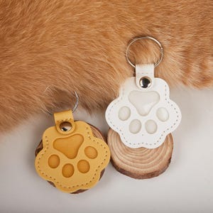 Puede incluir: Dos llaveros con forma de pata. Uno es de cuero amarillo, el otro de cuero blanco. Ambos tienen un llavero plateado y están unidos a una pequeña rodaja de madera. El fondo es una textura marrón claro y peluda.