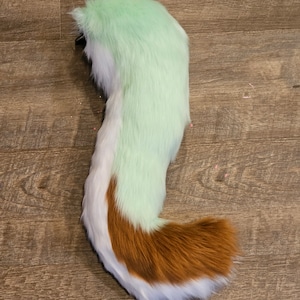 May include: A fluffy green, white, and brown faux fur tail with a white tip. The tail is attached to a black clip.