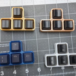 May include: Four T-shaped macro keypads with black keycaps. The top left is gold, the top right is copper, the bottom left is blue, and the bottom right is white. Each keypad holds four keys with various printed symbols and labels like F5, M4, and M6.