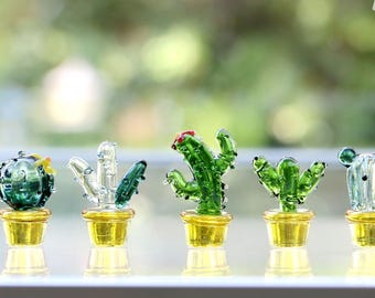 Może przedstawiać: Pięć miniaturowych szklanych rzeźb kaktusów w różnych kształtach i kolorach, każda w małym, złotym pojemniku. Kaktusy są w odcieniach zieleni, przezroczystości i błękitu, ze szczegółami takimi jak kwiaty. Tło jest rozmyte.