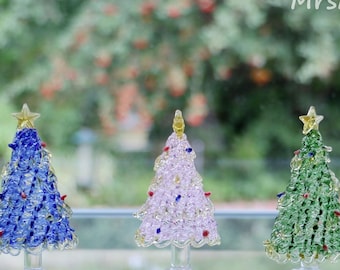 Peut inclure: Trois sapins de Noël miniatures en verre, un bleu, un rose et un vert, avec des étoiles dorées en guise de sommet. Les sapins sont posés sur une surface en verre transparent.