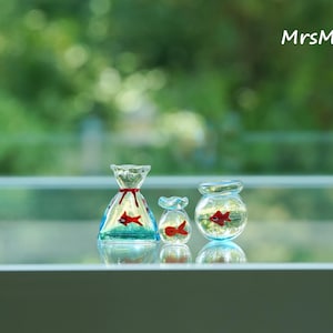 May include: Three miniature glass fish bowls, each containing a red fish. The bowls vary in shape: a bag, a small jar, and a round bowl. The bag-shaped bowl has a red ribbon. The background is a blurred green.