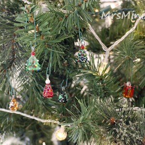 Op de afbeelding: Een close-up van een kerstboom versierd met kleine glazen ornamenten in de vorm van kerstboompjes. De ornamenten zijn in verschillende kleuren, waaronder rood, groen en bruin. De ornamenten hangen aan de takken van de boom met een doorzichtige draad.
