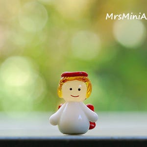 May include: A small, glass angel figurine with a white body, yellow hair, and a red halo. The angel has a smiling face and small red wings. The background is a blurred green and white bokeh effect. The text "MrsMiniArts" is in the upper right corner.