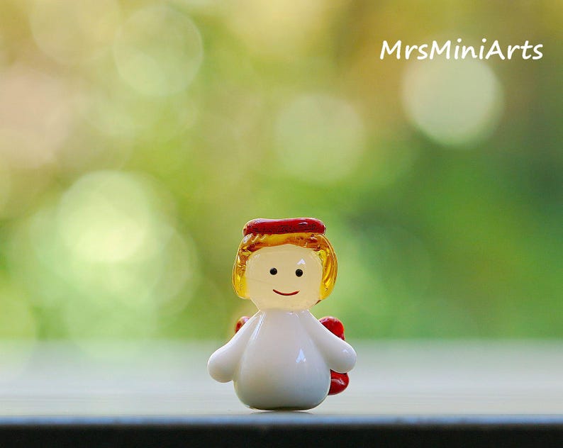 May include: A small, glass angel figurine with a white body, yellow hair, and a red halo. The angel has a smiling face and small red wings. The background is a blurred green and white bokeh effect. The text "MrsMiniArts" is in the upper right corner.